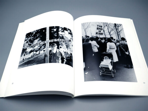 Detail of the "Sign Language" exhibit catalogue, showing the book open to photos of a bearded man holding a large wooden cross that reads "VOTE 4 JESUS," and another photo of a crowd of demonstrators, facing away from the camera, with a sign reading "THE END IS AT HAND" and a sleeping infant in a baby carriage.